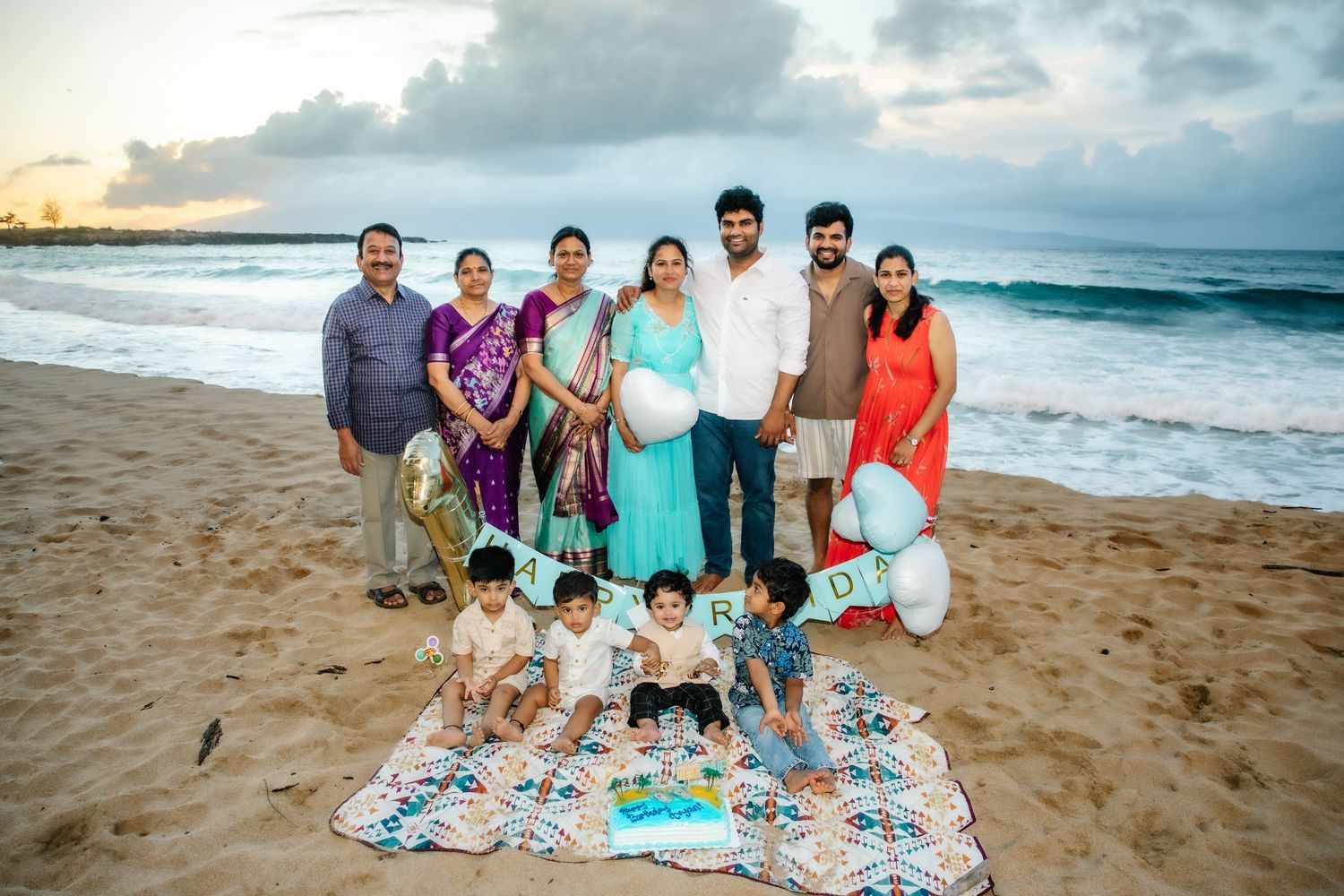 Indian family celebrating on a sandy beach at sunset with balloons, children seated on a blanket with decorative letters.