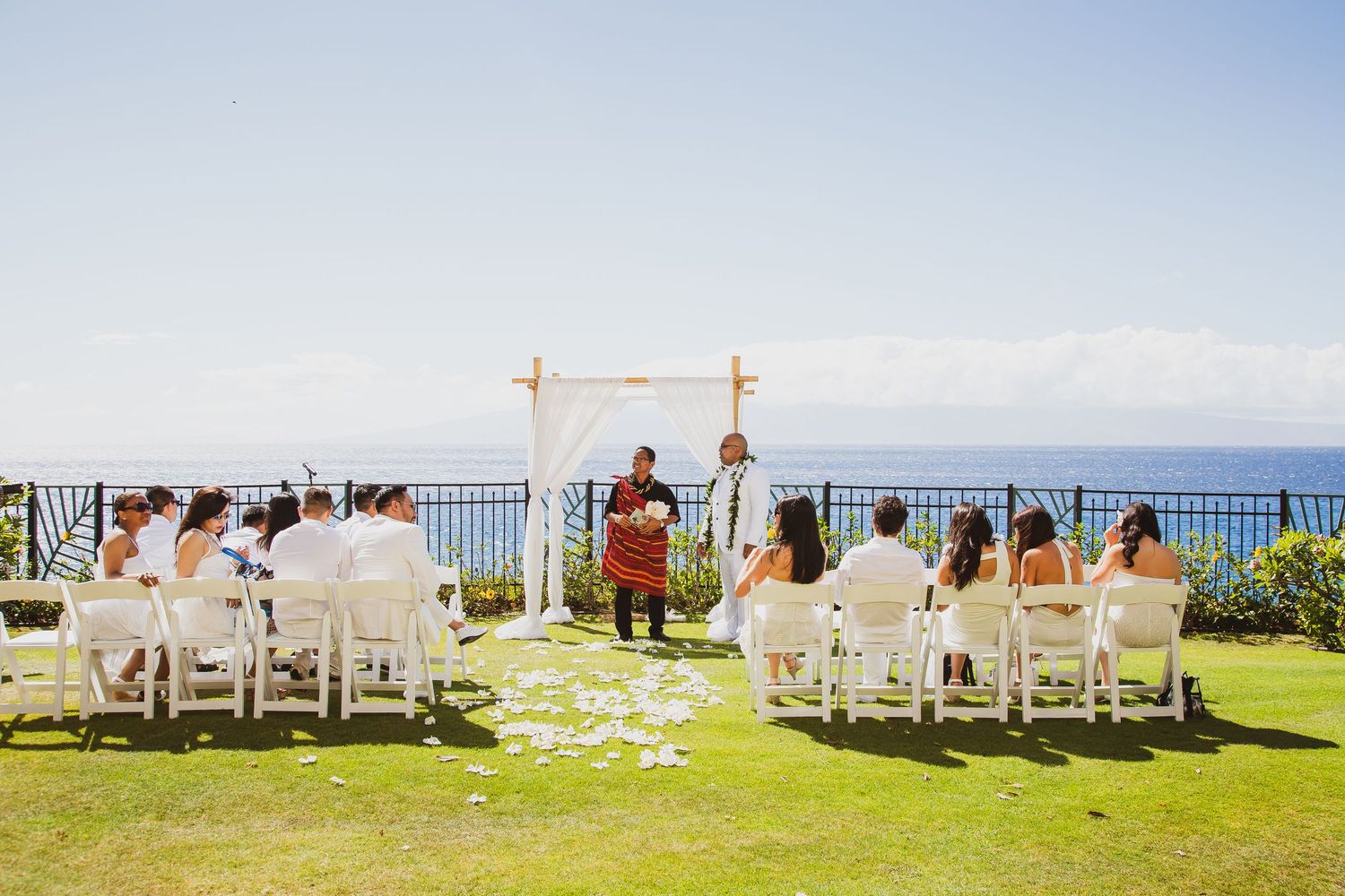 Outdoor wedding ceremony on a lush green lawn overlooking the ocean, with guests seated in white chairs under a bamboo arch.