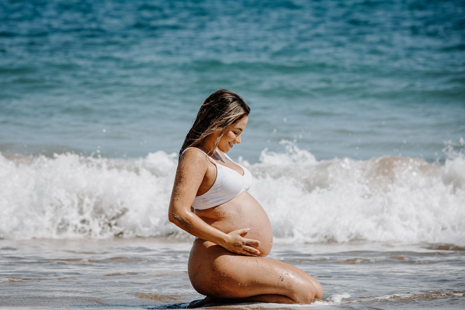 Pregnant woman sitting in ocean waves during sunset maternity photoshoot.
