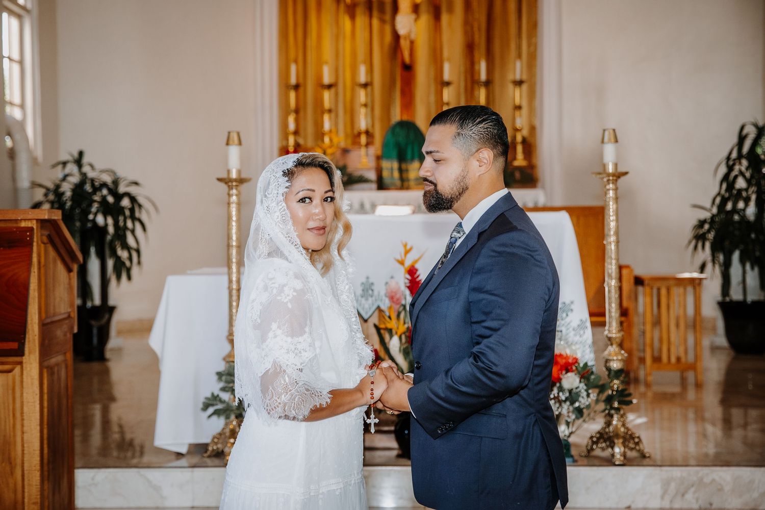 Bride and groom exchange vows at a church altar, bride wearing lace veil and groom in navy suit.