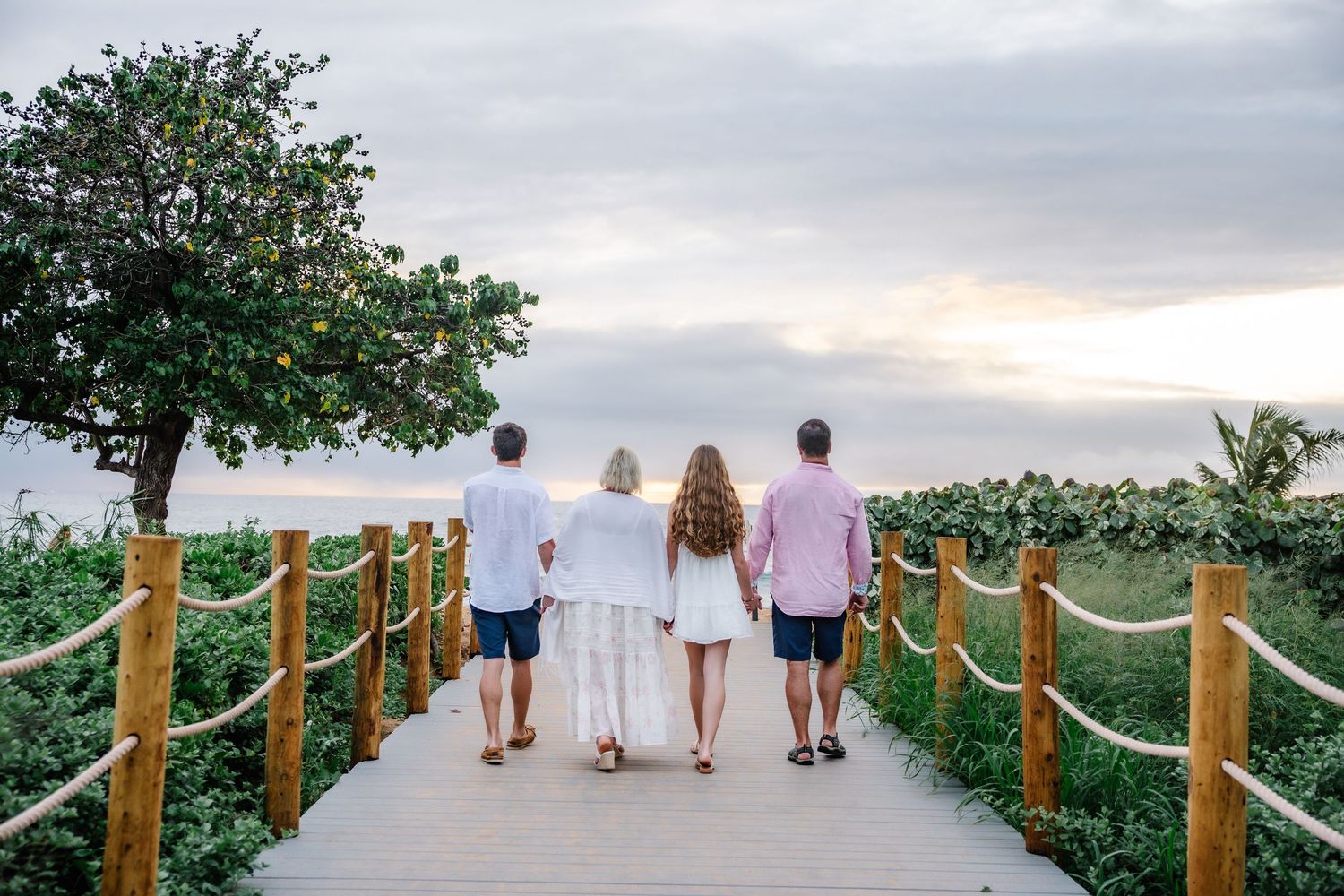Family of four walking on wooden boardwalk toward ocean sunset surrounded by tropical greenery.