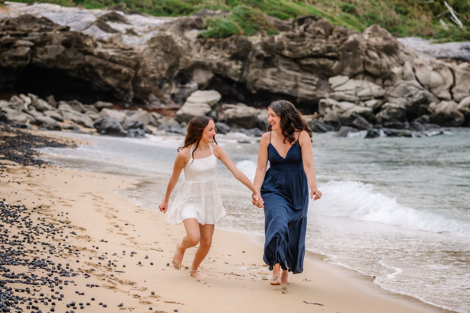 Two women in dresses hold hands walking along a sandy beach with rocky cliffs in the background.