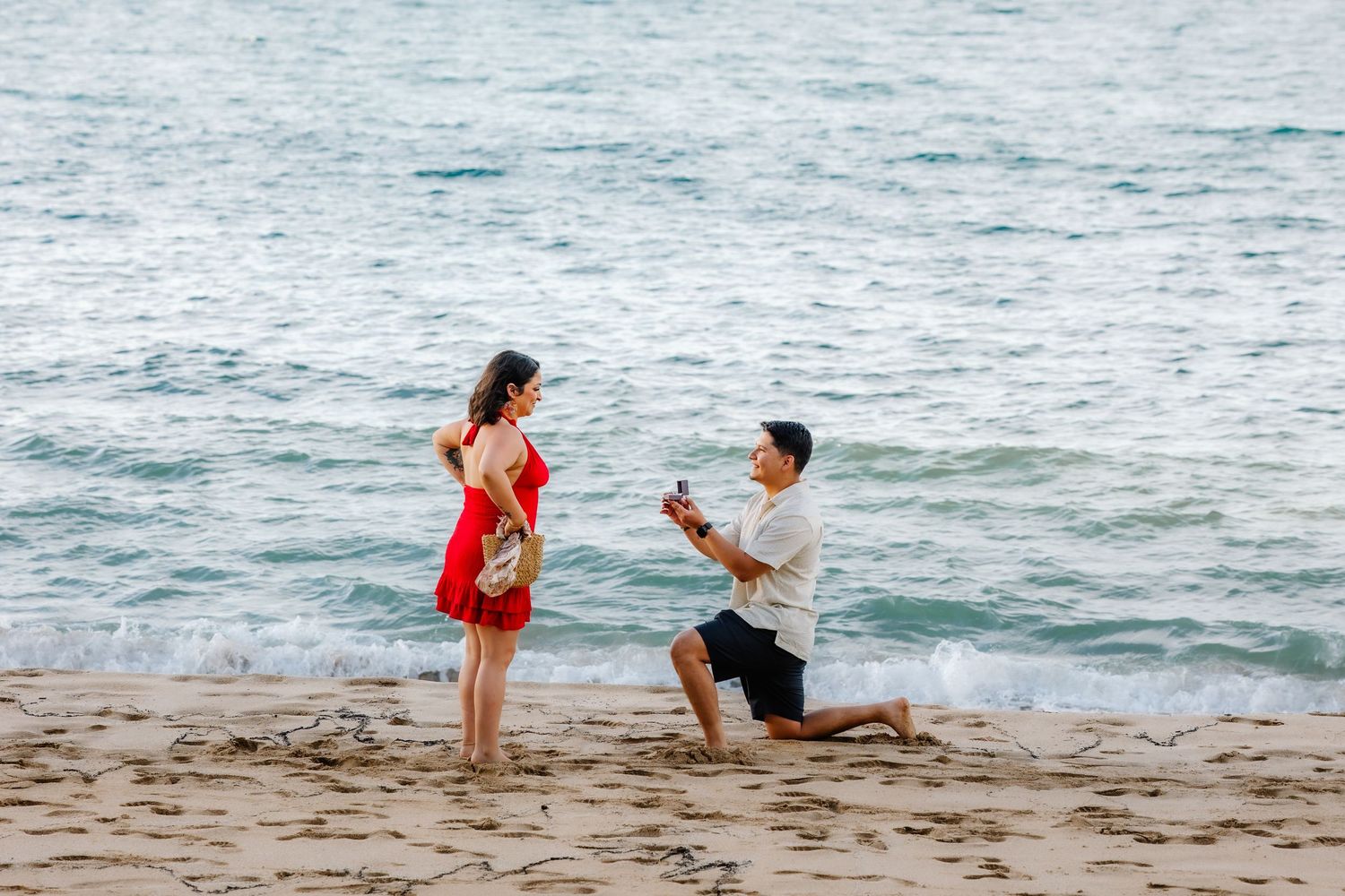 Man proposes to woman on sandy beach, kneeling with ring box while she stands in red dress by ocean waves.