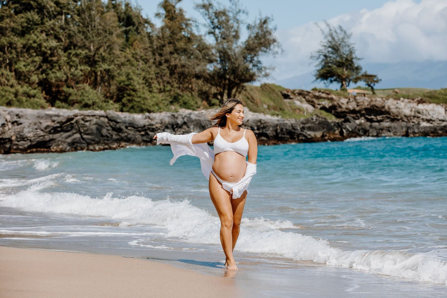 A maternity beach photo with a person in white walking along turquoise waters and rocky coastline.