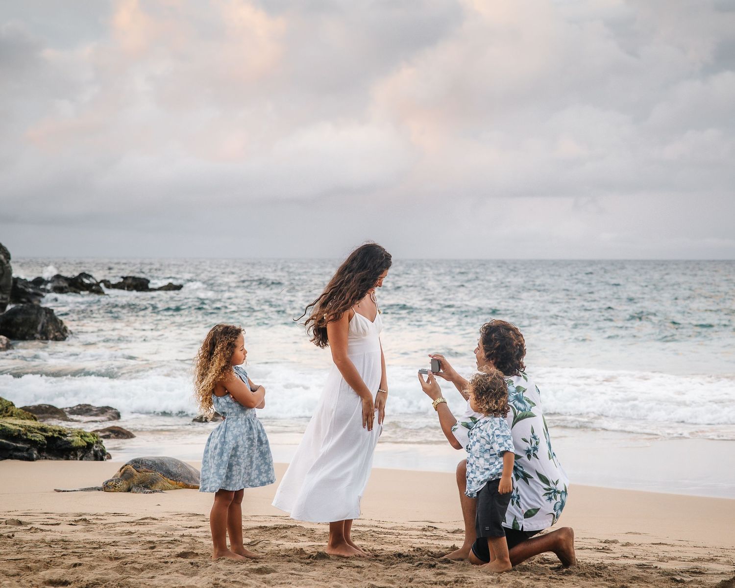 Family enjoying a sunset beach photoshoot in coordinated blue and white outfits on a sandy Hawaiian shore.