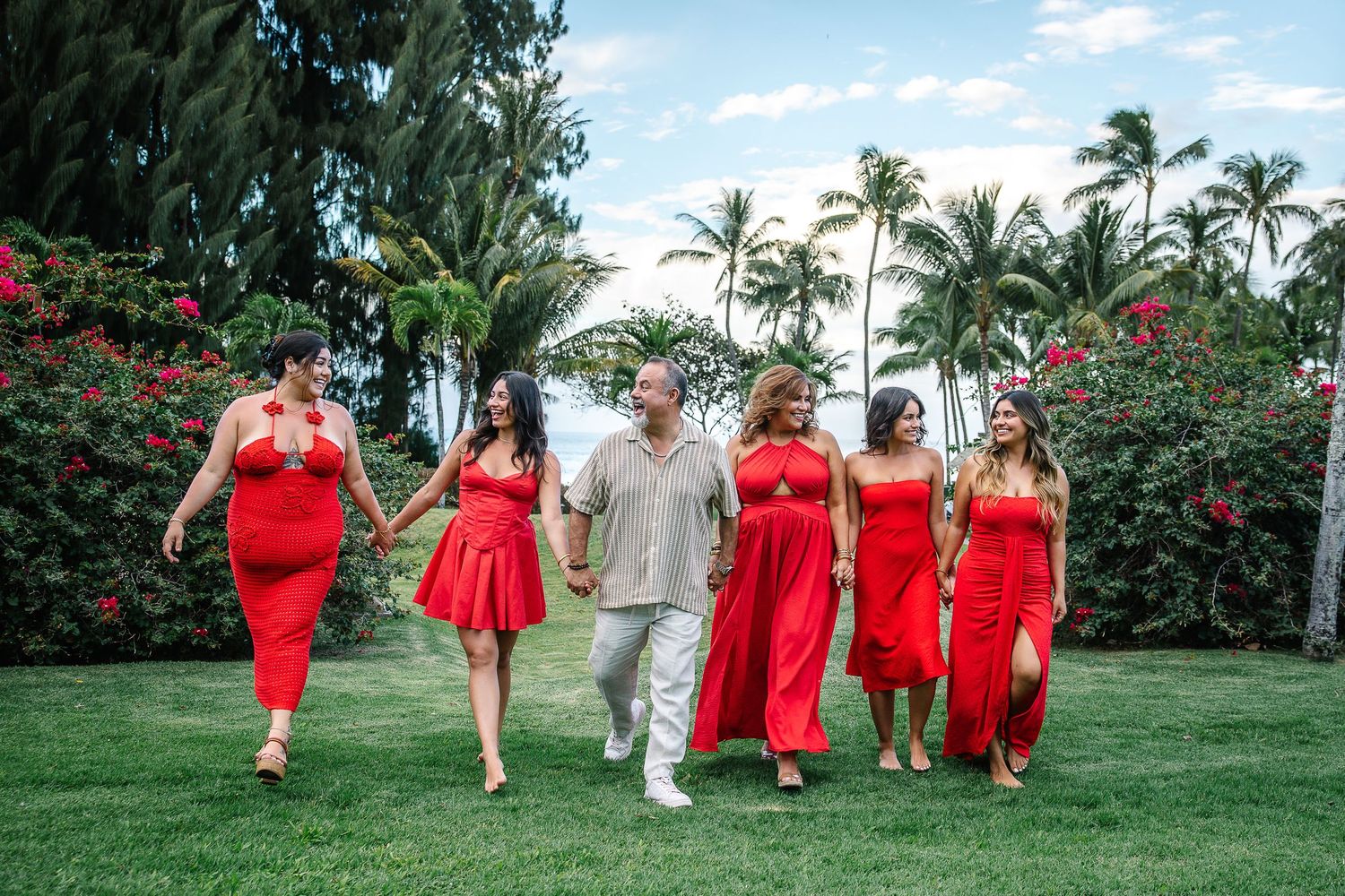 Wedding group in red and white attire poses on grass with tropical landscaping.