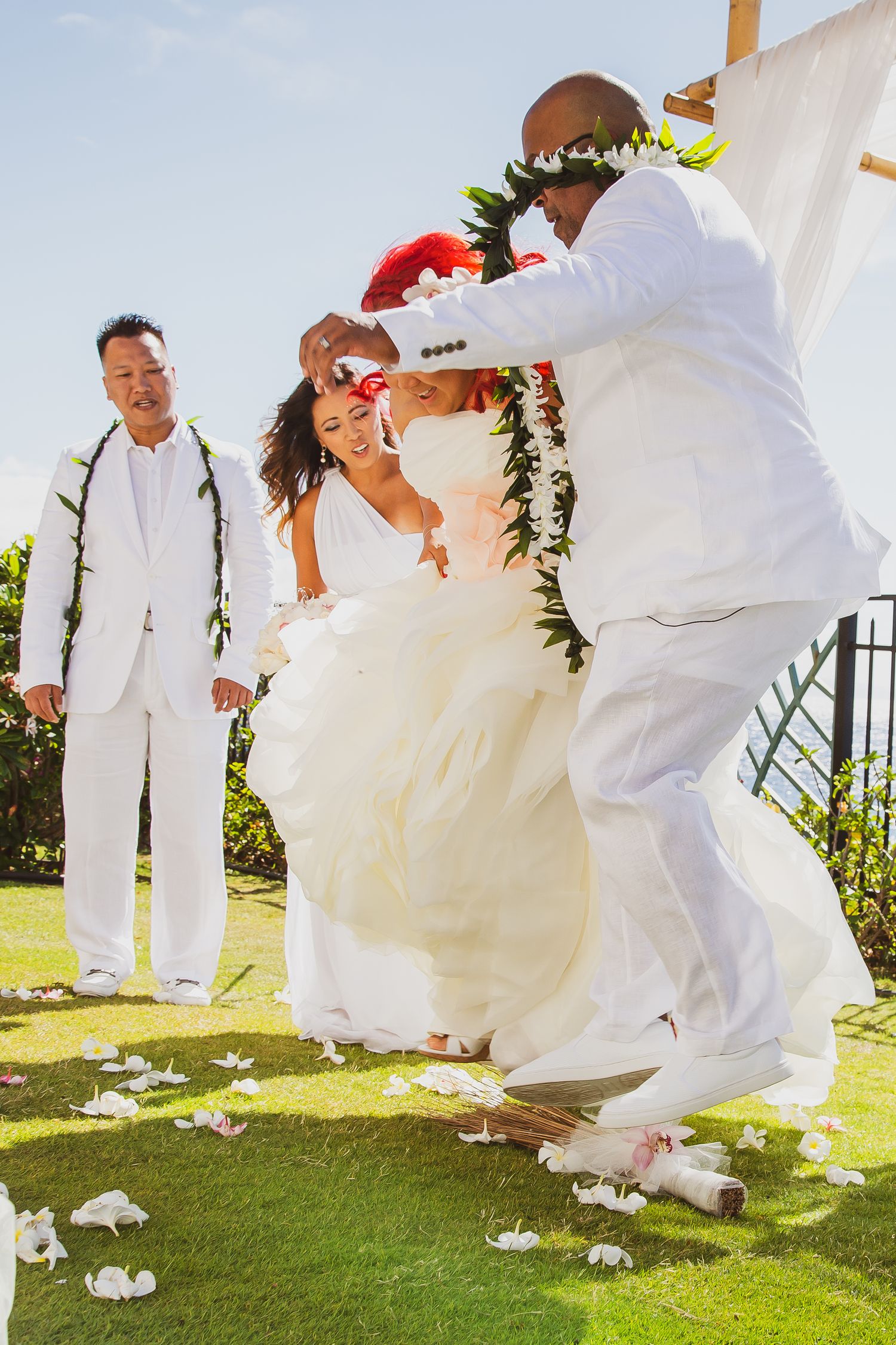 Bride and groom in white attire celebrate at outdoor Hawaiian wedding ceremony with leis and flower petals on grass.