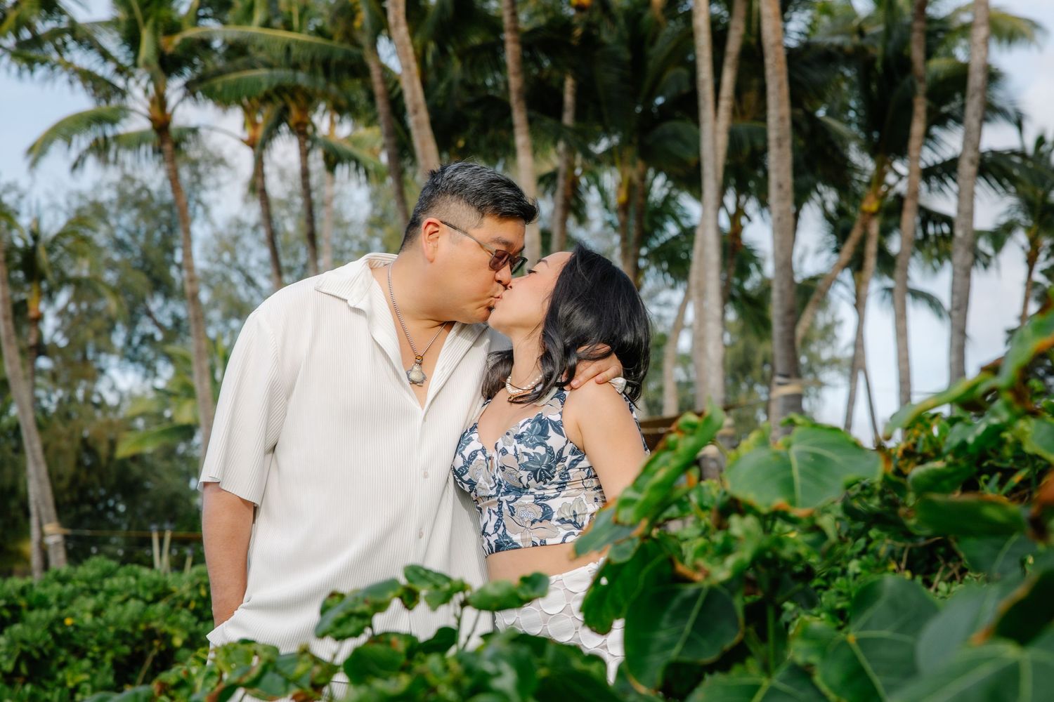 Couple kissing among tropical greenery with palm trees in background, romantic outdoor photo session.