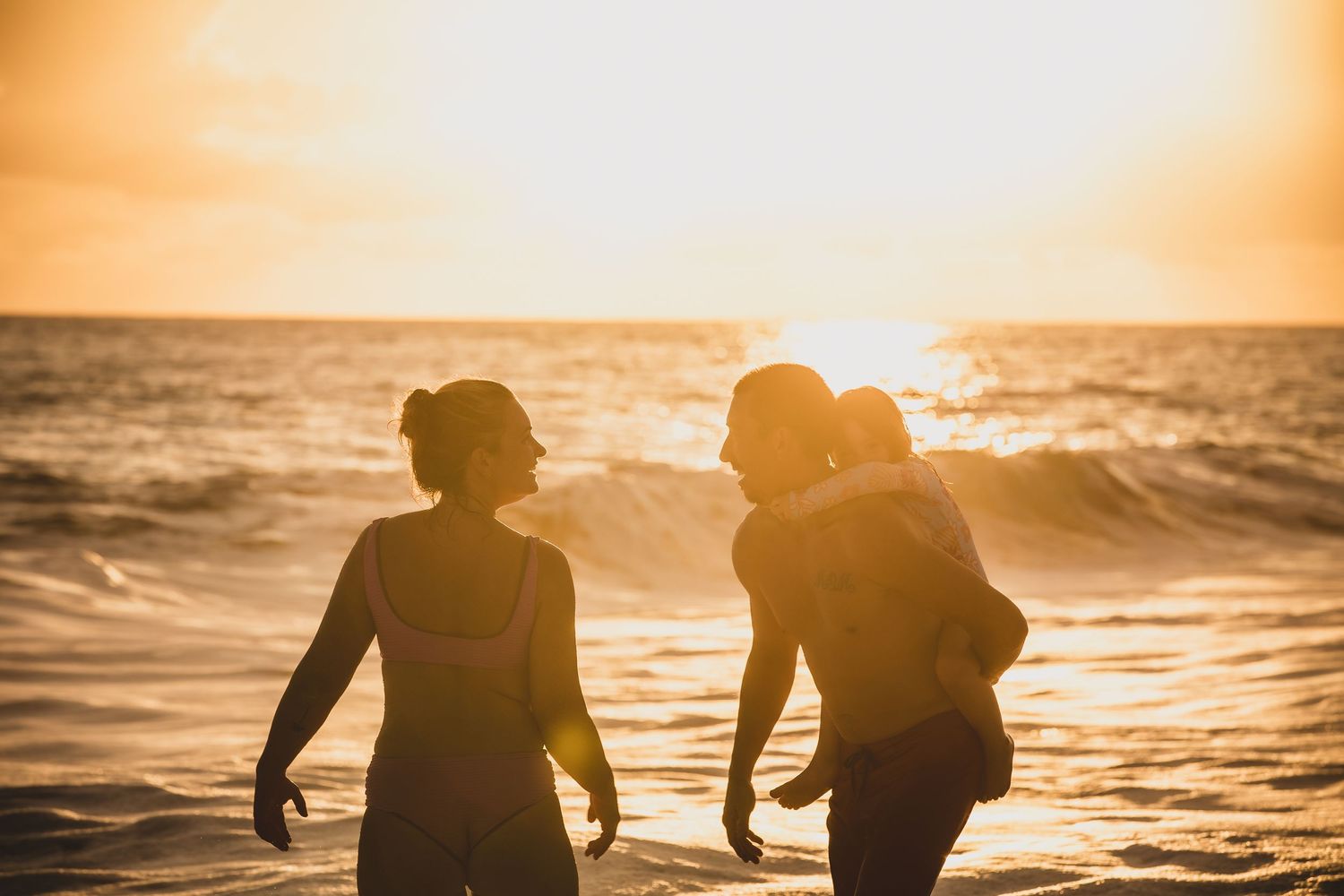 Two women and a child silhouetted against a golden sunset on a beach with ocean waves in the background.