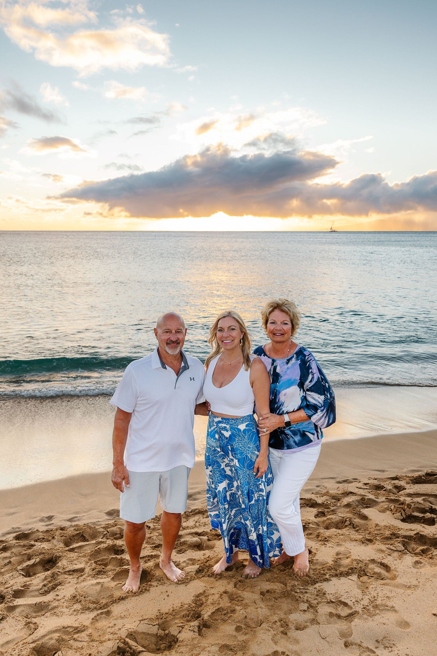 Three people in coordinating blue and white outfits pose together on a sandy beach at sunset with ocean waves behind them.
