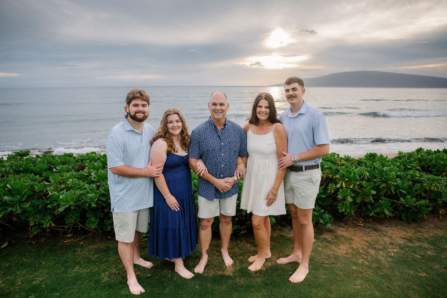 A group of people stand together on a beach at sunset with ocean waves and cloudy skies in the background.