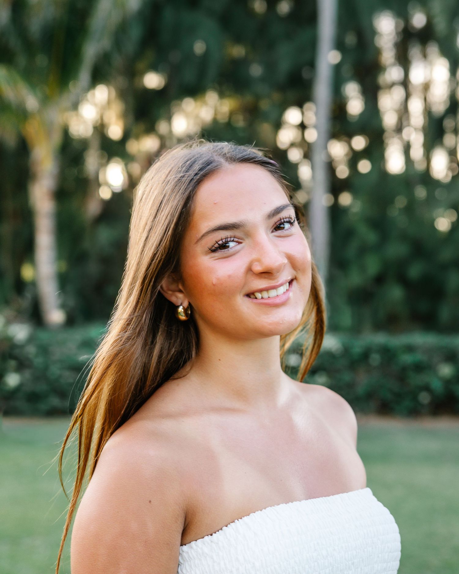 Portrait of a young woman in a white dress smiling outdoors with palm trees in background.