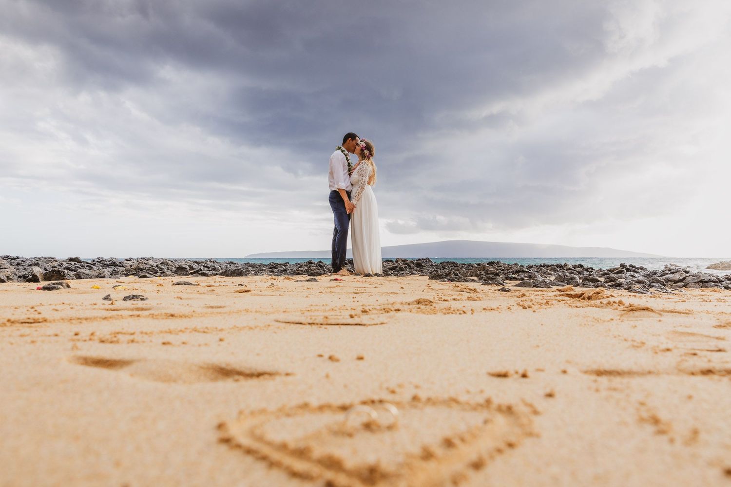 Couple enjoying a romantic moment on a scenic beach with dramatic cloudy skies and rocky coastline.