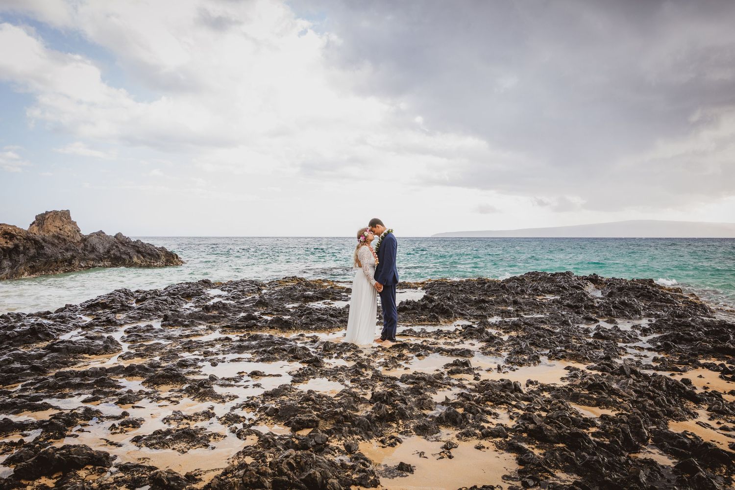 Bride and groom share a romantic kiss on a rocky Hawaiian beach with turquoise ocean waters and cloudy skies.