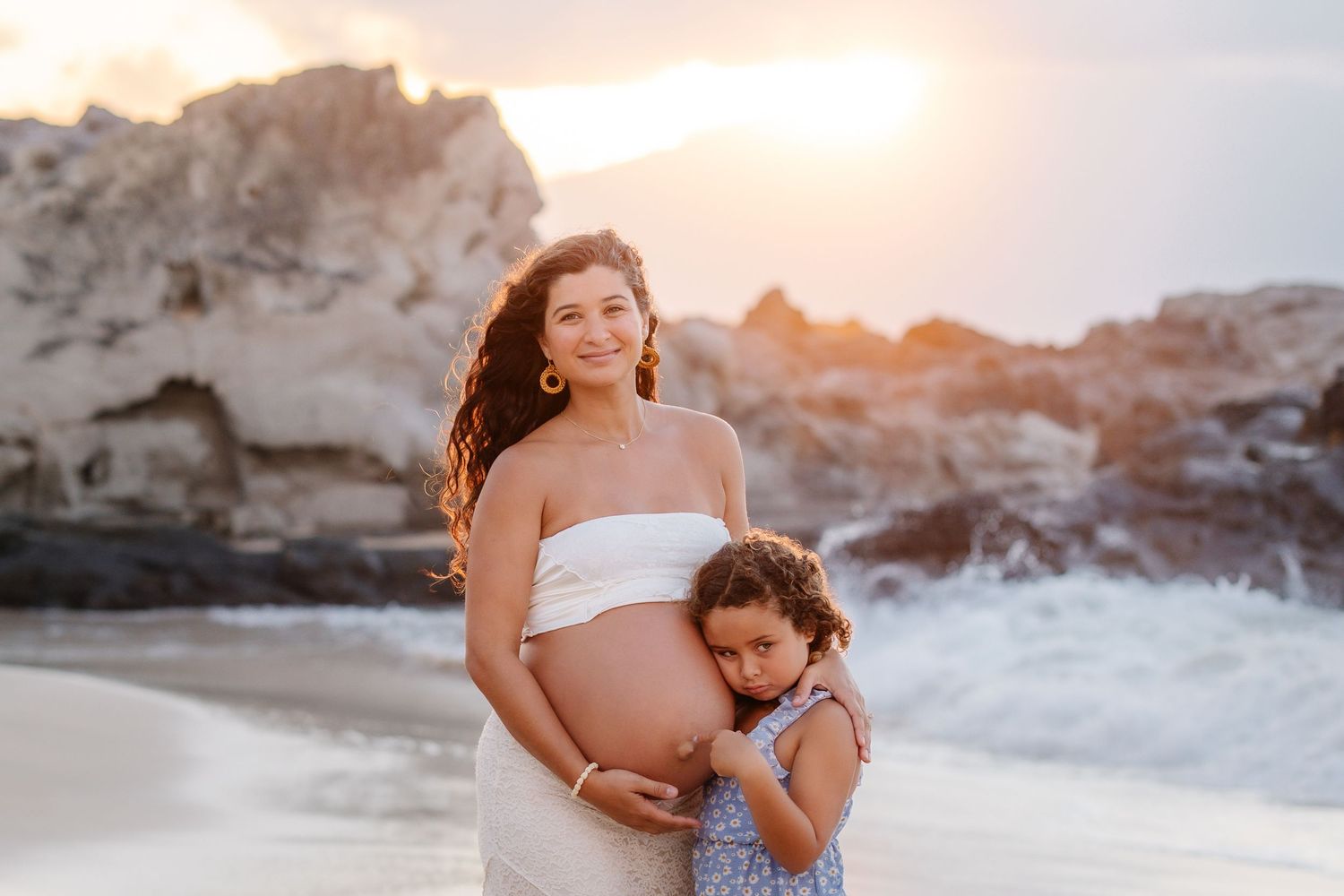 Pregnant woman in white dress hugging young daughter on beach at golden sunset with rocky shore backdrop.