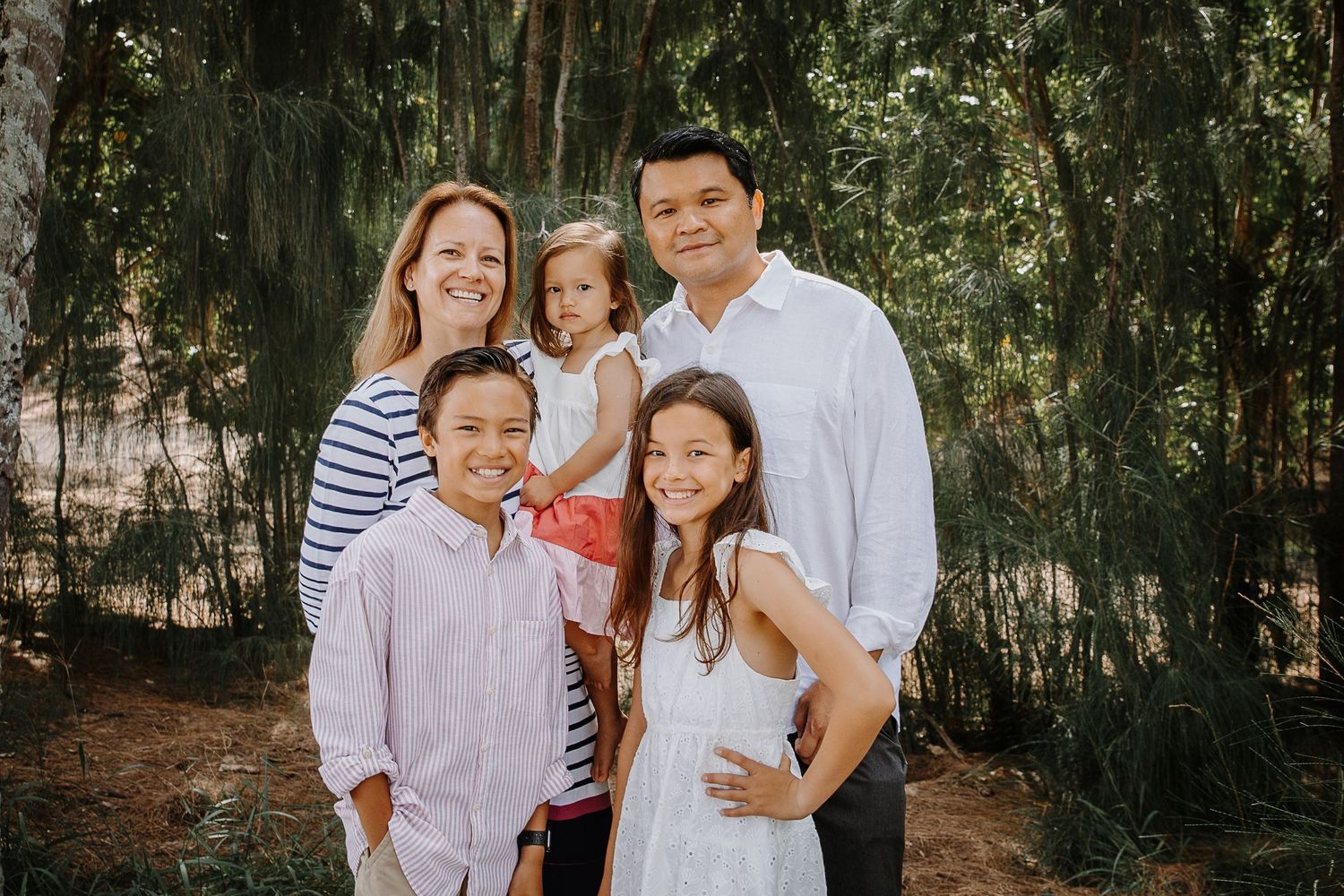 A group poses together outdoors in white clothing among trees and natural greenery for a family photo session.