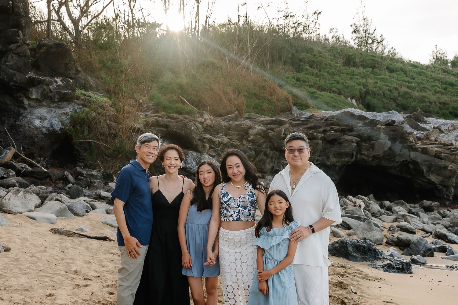 Family of six posing together on a sandy beach with rocky cliffs and sunlit trees in the background.
