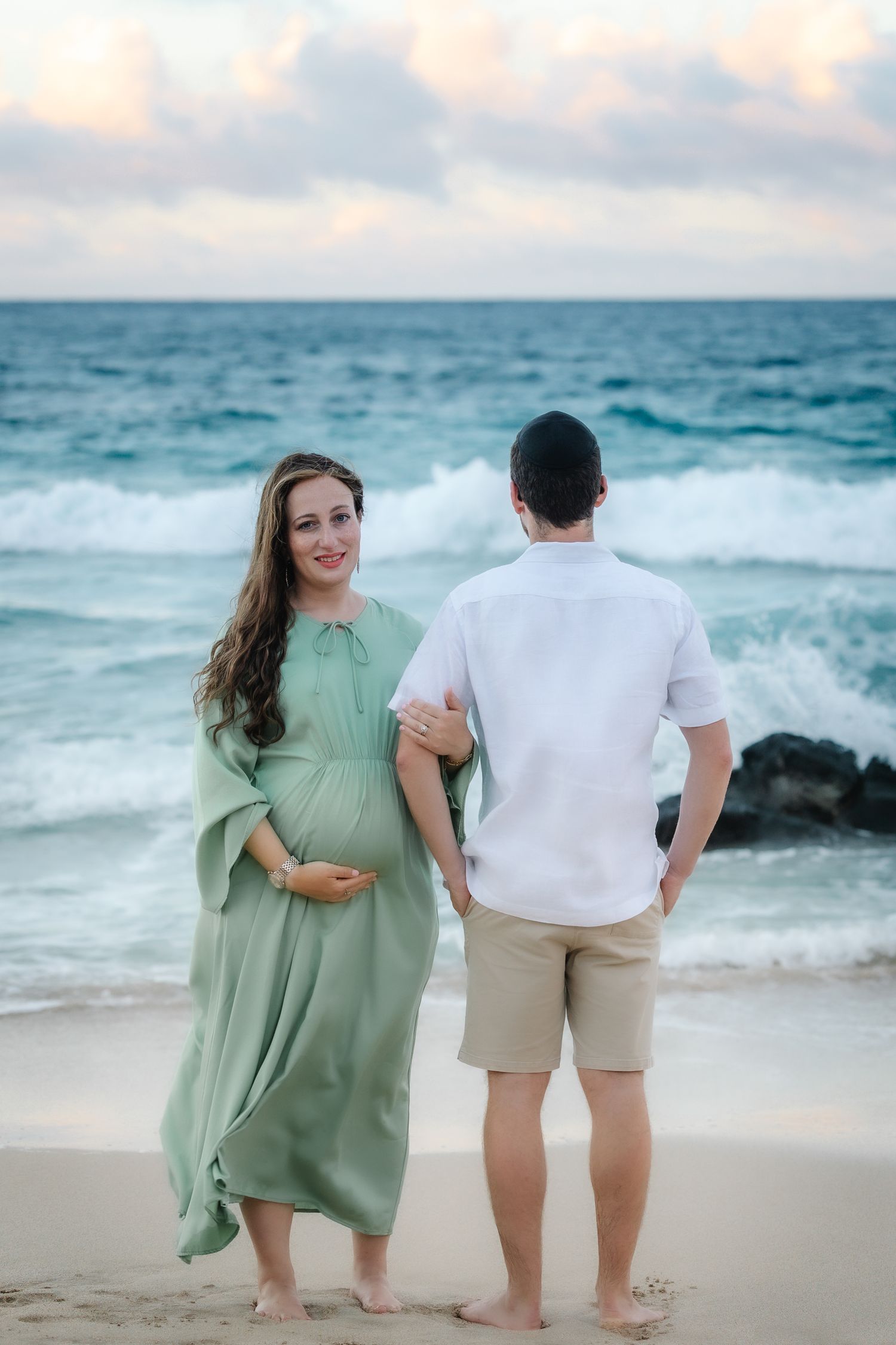 Pregnant woman in green dress with partner on beach at sunset, waves crashing behind them.