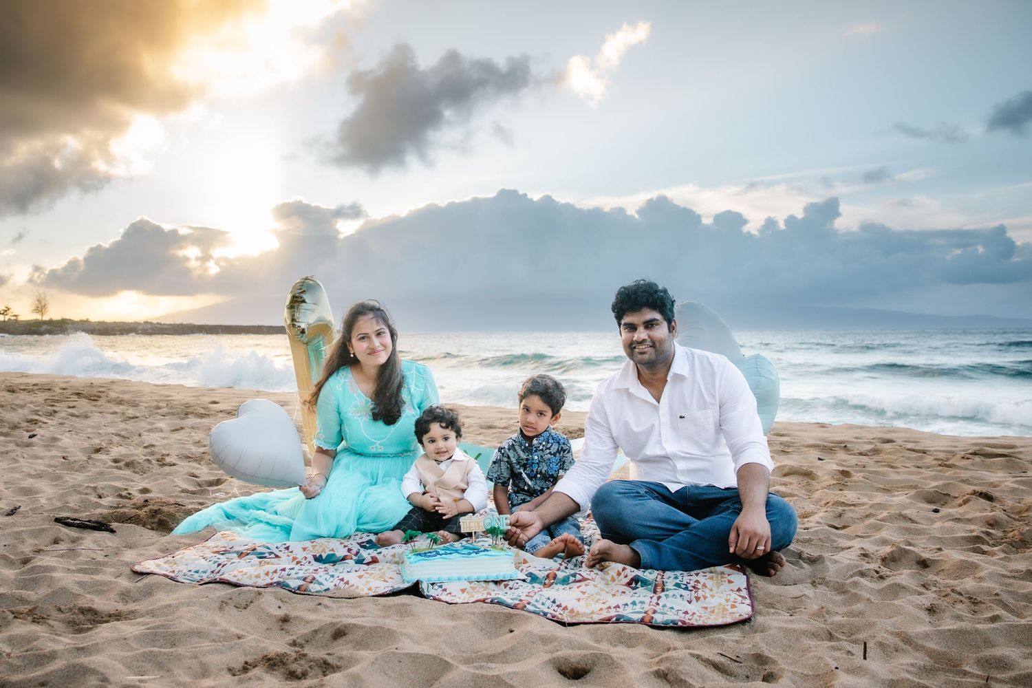 Family of four sitting on sandy beach at sunset, two parents with toddlers on colorful blanket by ocean waves.