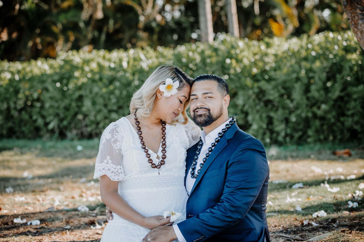 Couple in wedding attire smiling outdoors, bride in white lace dress with flower in hair, groom in navy suit.