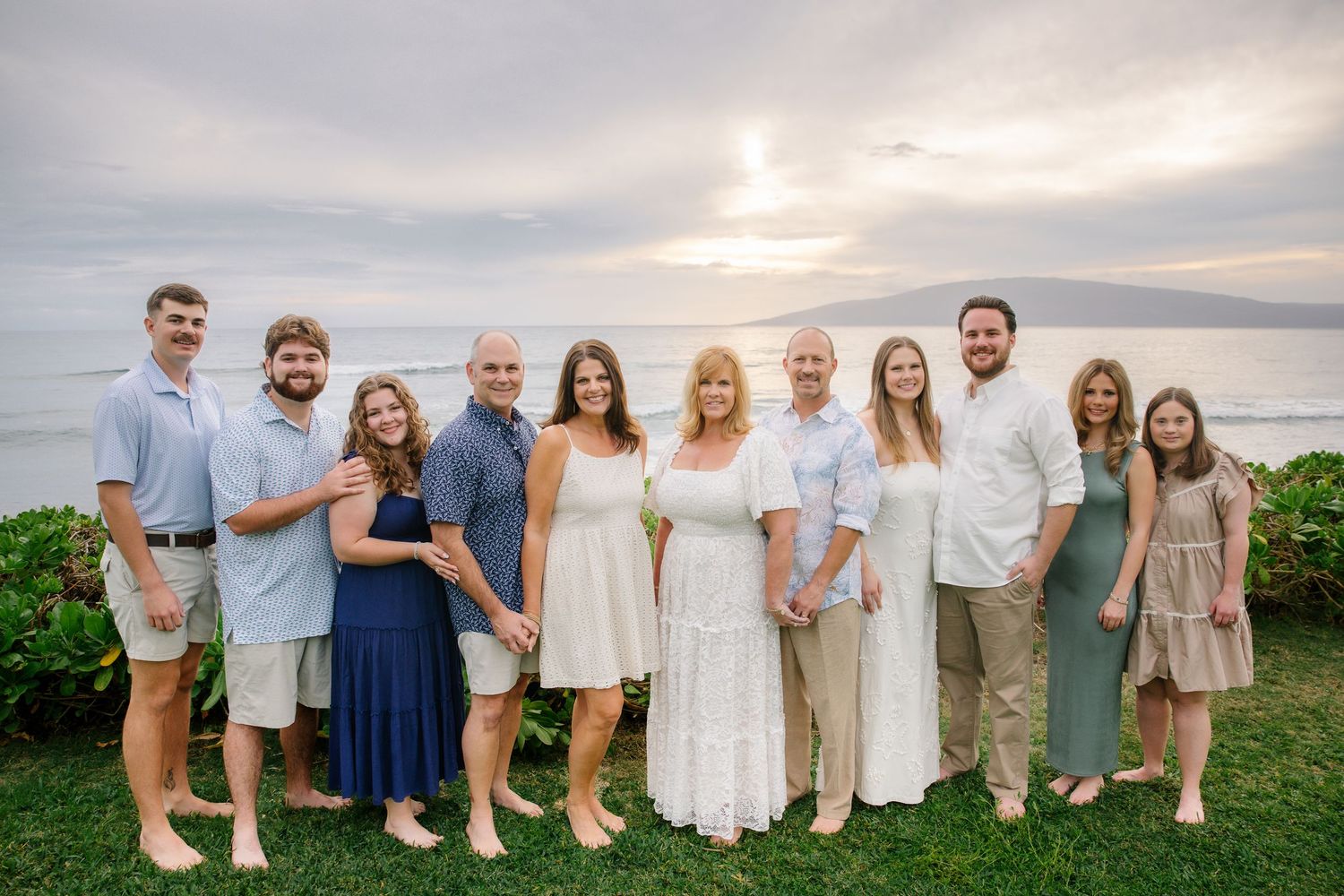 Family group wearing coordinated white and blue outfits poses together at sunset on a grassy oceanfront location.