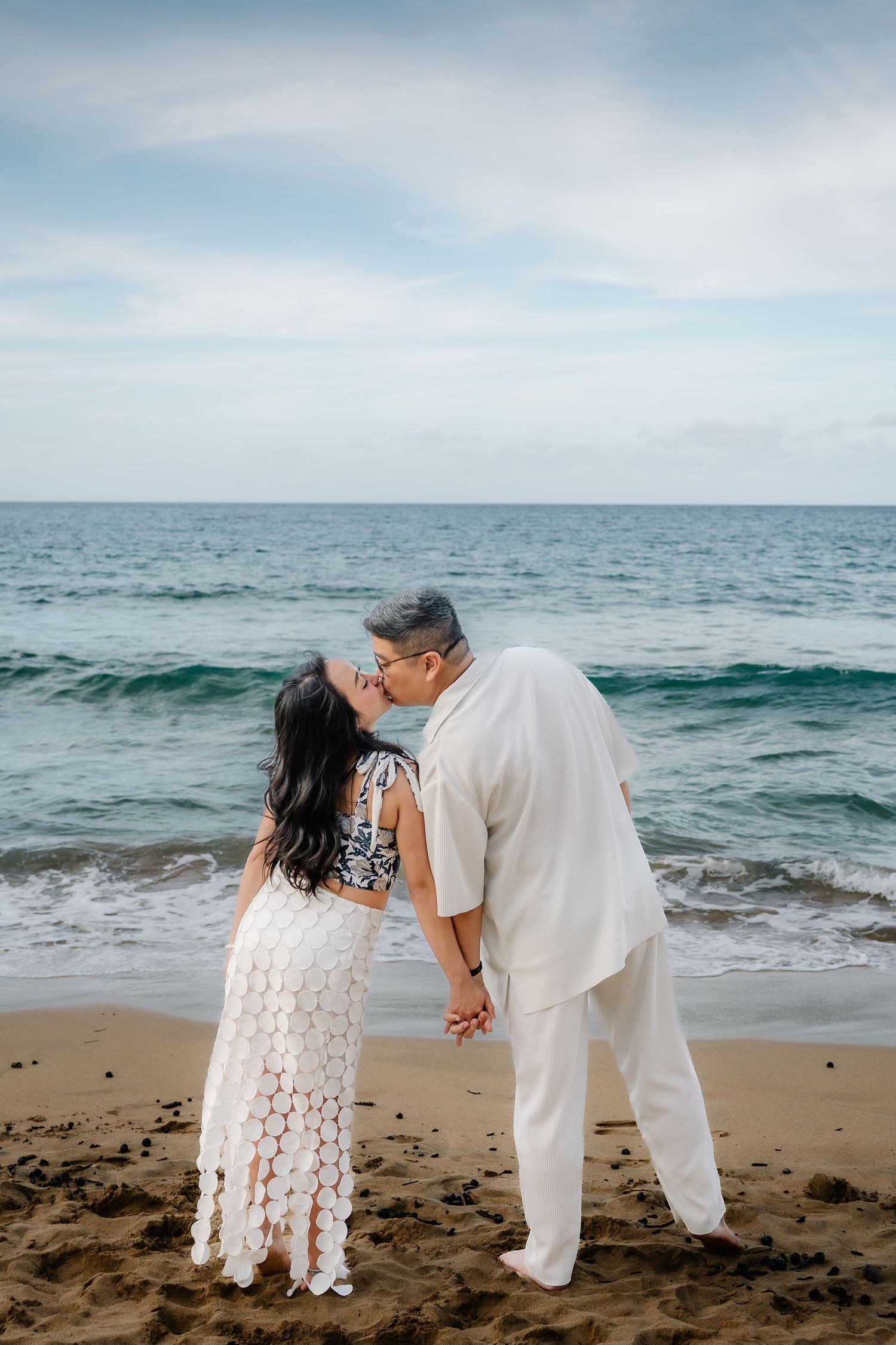 Couple sharing a romantic kiss on a sandy beach, woman in white lace dress, man in white outfit, ocean waves behind them.