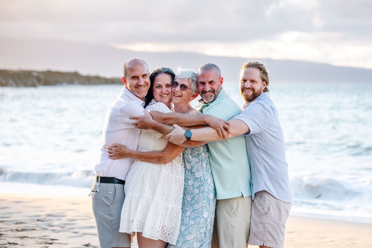 Five adults hugging and smiling together on a sandy beach with ocean waves and cloudy sky in the background.
