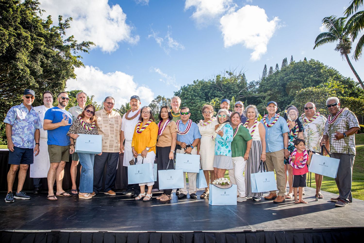 Large family gathering outdoors with tropical landscaping and blue sky background.
