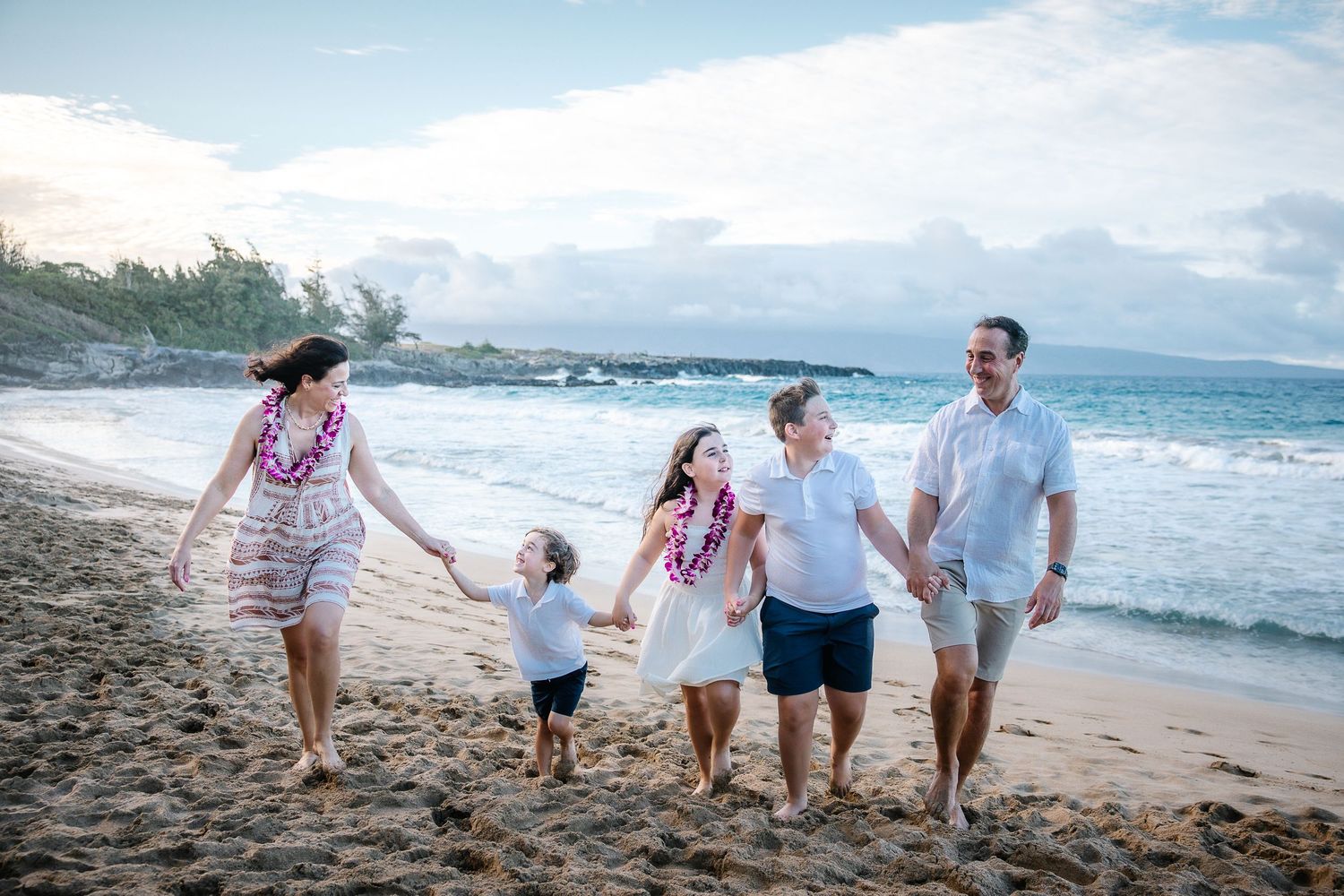 Family holding hands while walking along a scenic beach with waves and palm trees.