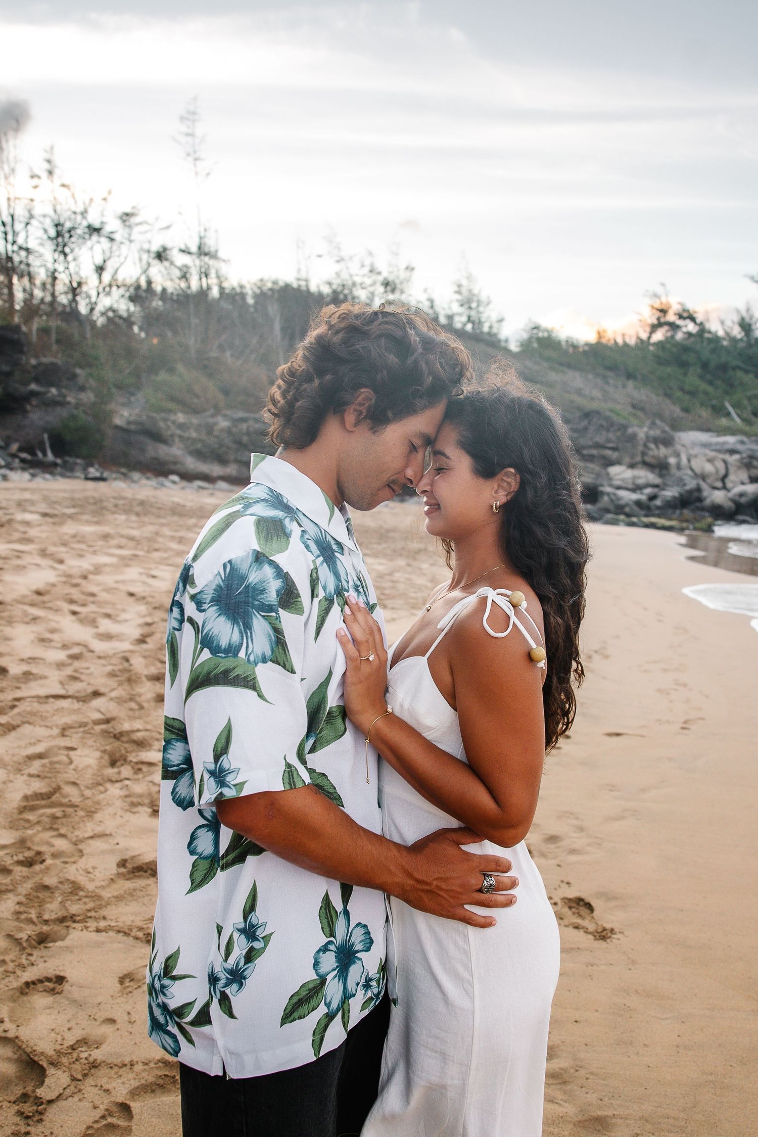 A couple embraces tenderly on a sandy beach wearing coordinated white and tropical print outfits.