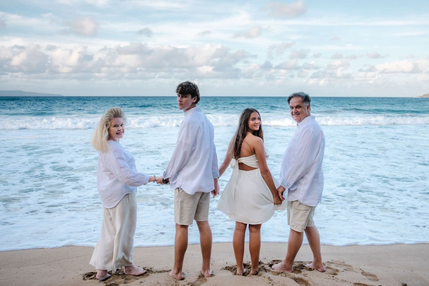 Family walking along beach shoreline in coordinated white outfits during sunset.
