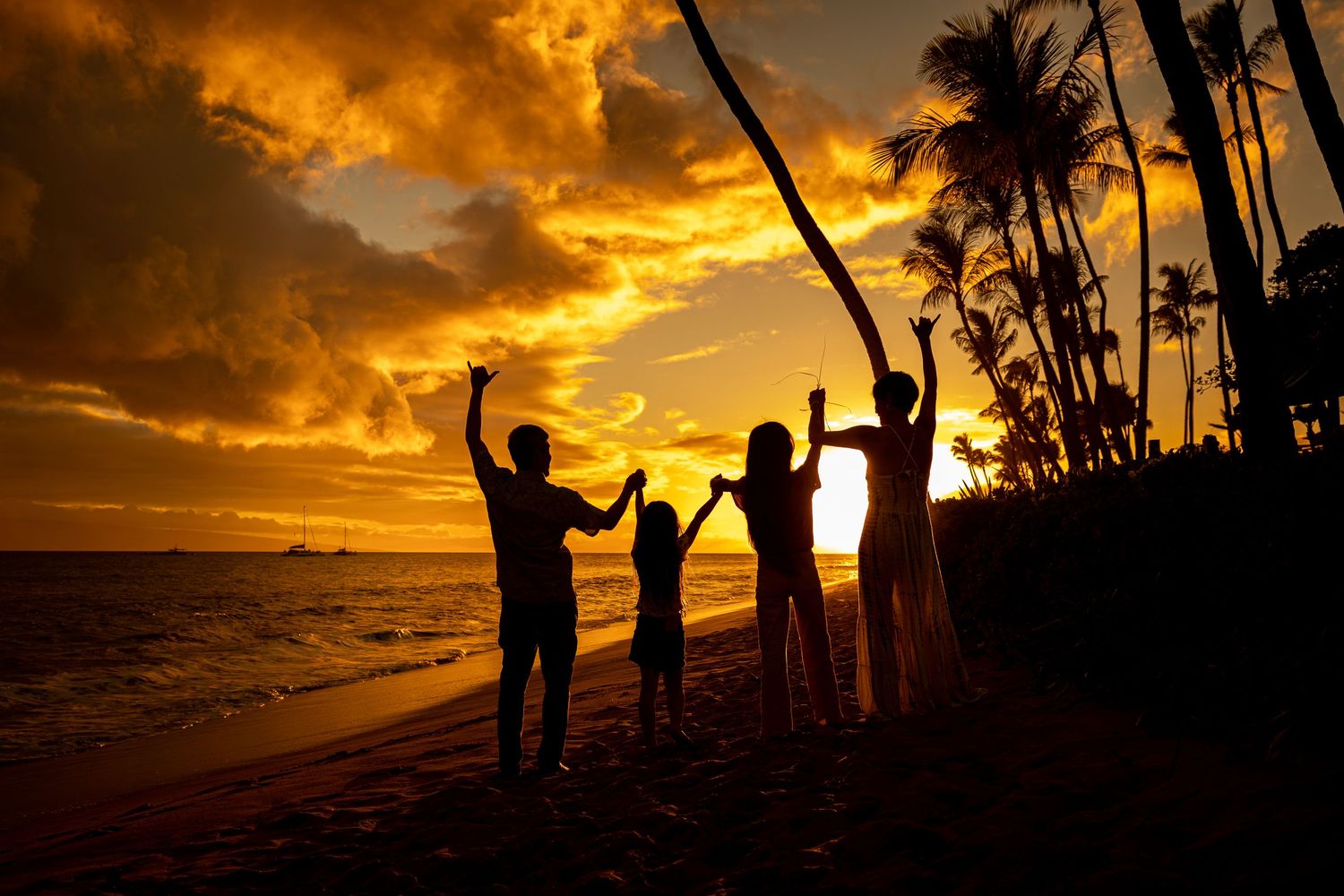 Family of four silhouetted against a vibrant tropical sunset on a beach, raising arms joyfully with palm trees nearby.