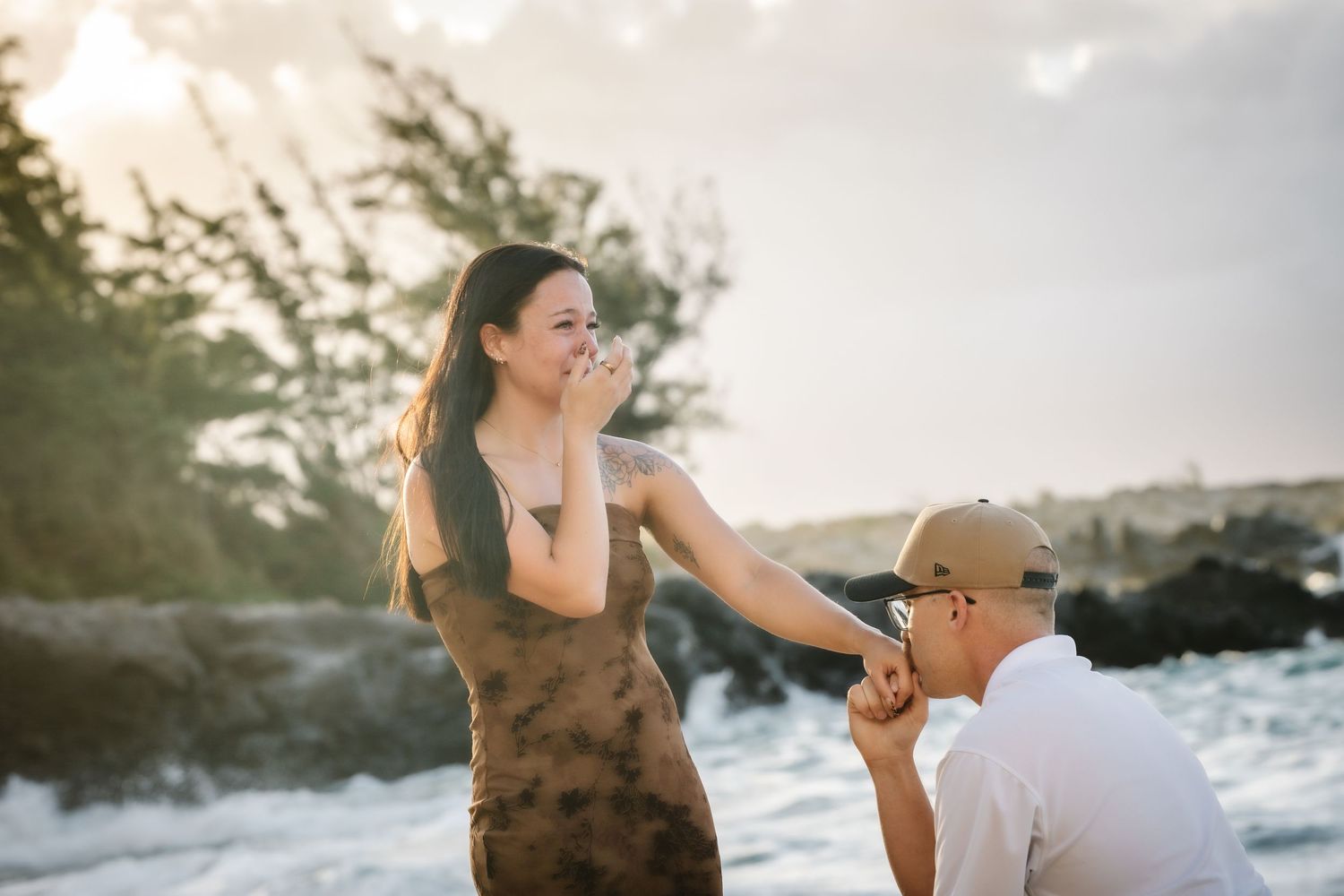 Man kneeling on beach proposing to surprised woman covering her mouth, ocean waves and golden sunset in background.
