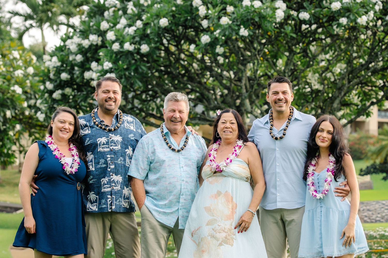 Group of six people wearing leis posing together outdoors in Hawaii with blooming white flowers in background.