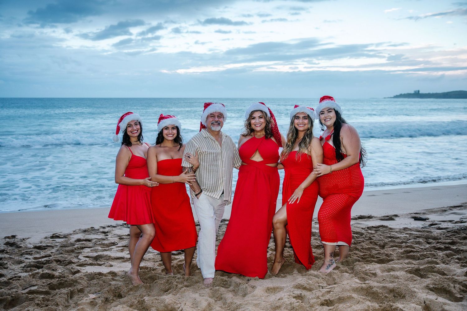 Festive beach wedding group wearing red dresses and Santa hats smiling by the ocean.