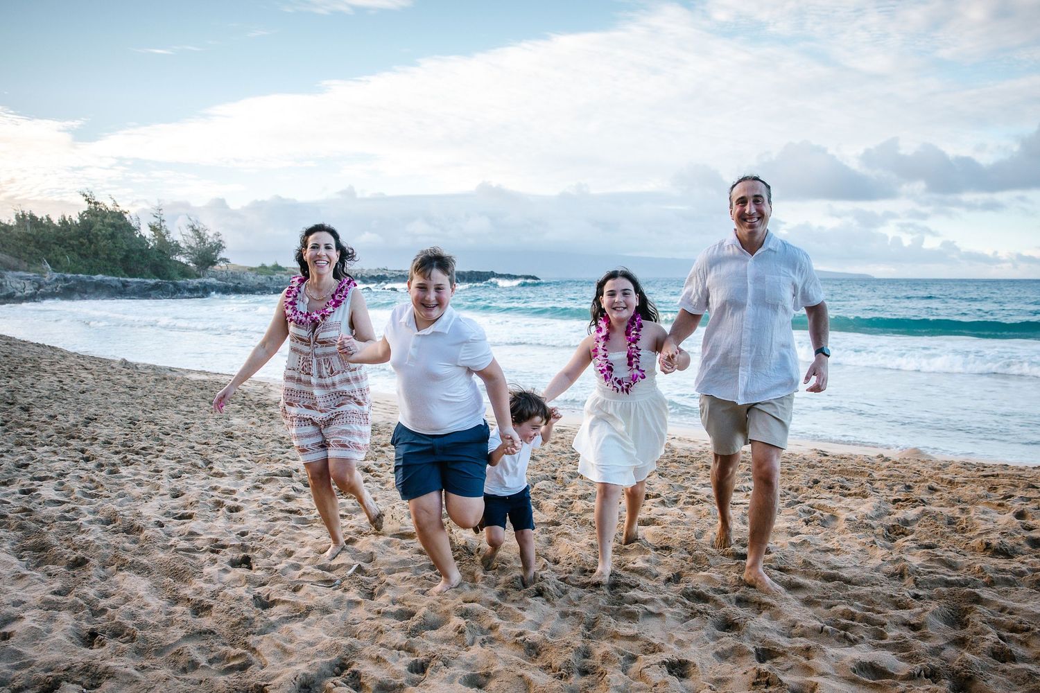 Family of four running and playing together on a tropical beach with waves in background.