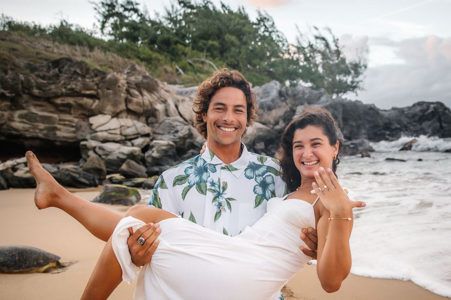 Couple shows off engagement ring during playful beach photoshoot with ocean views.