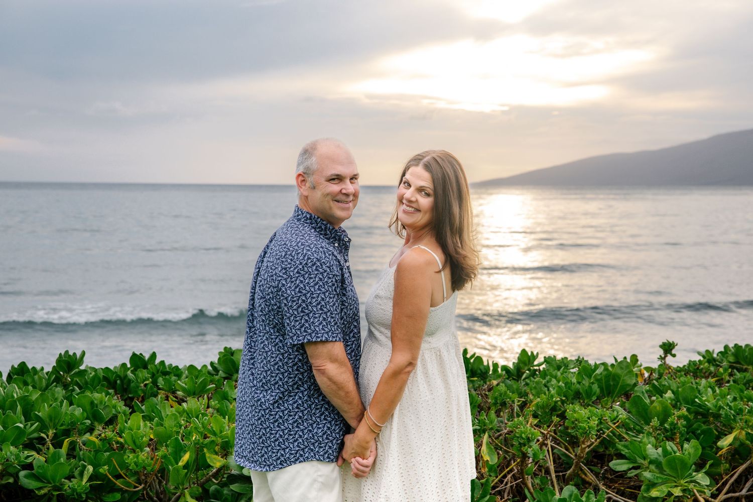 A couple embraces during sunset at a beach in Hawaii with lush tropical foliage in the foreground.