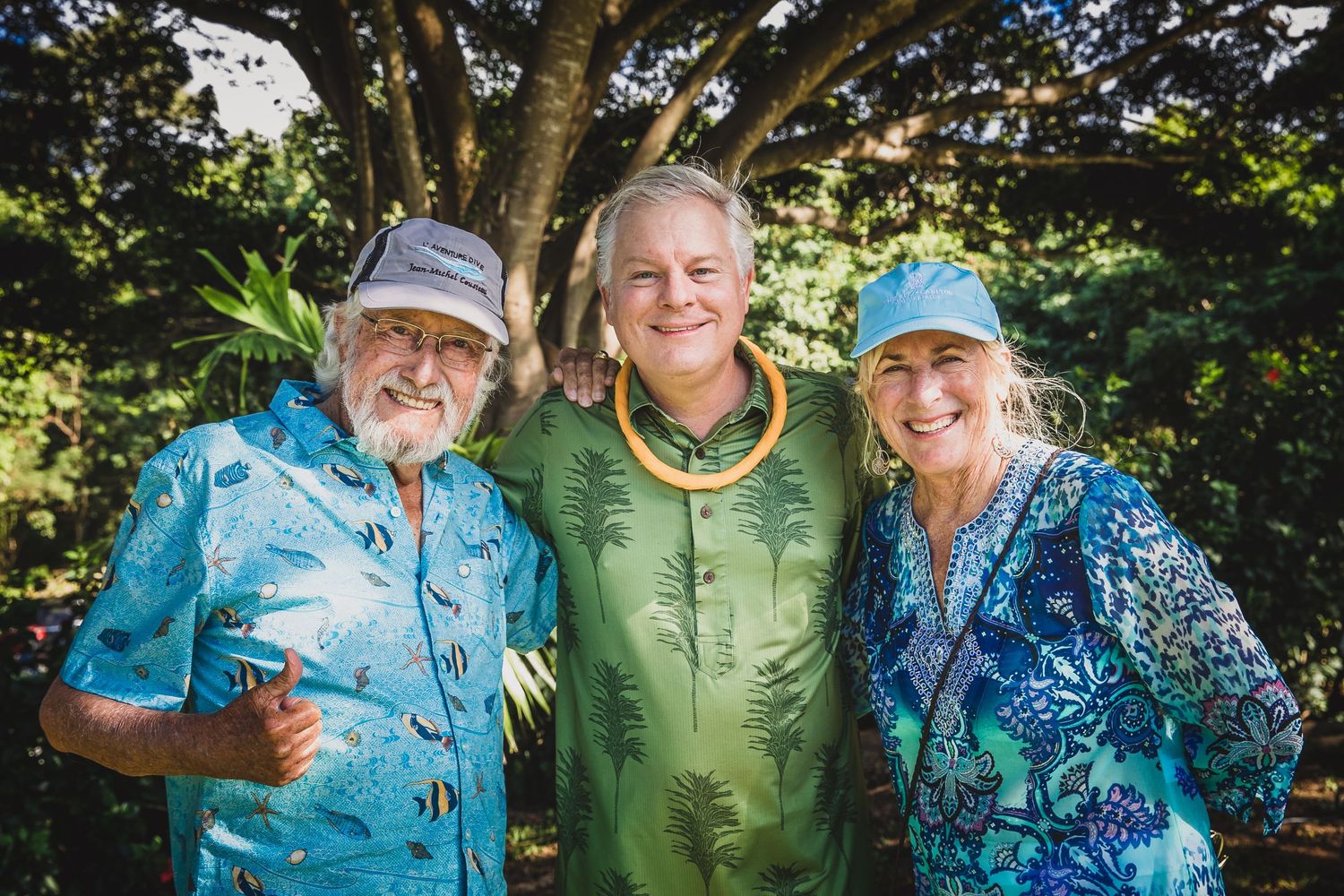Three people in matching blue tropical print shirts pose together under trees.