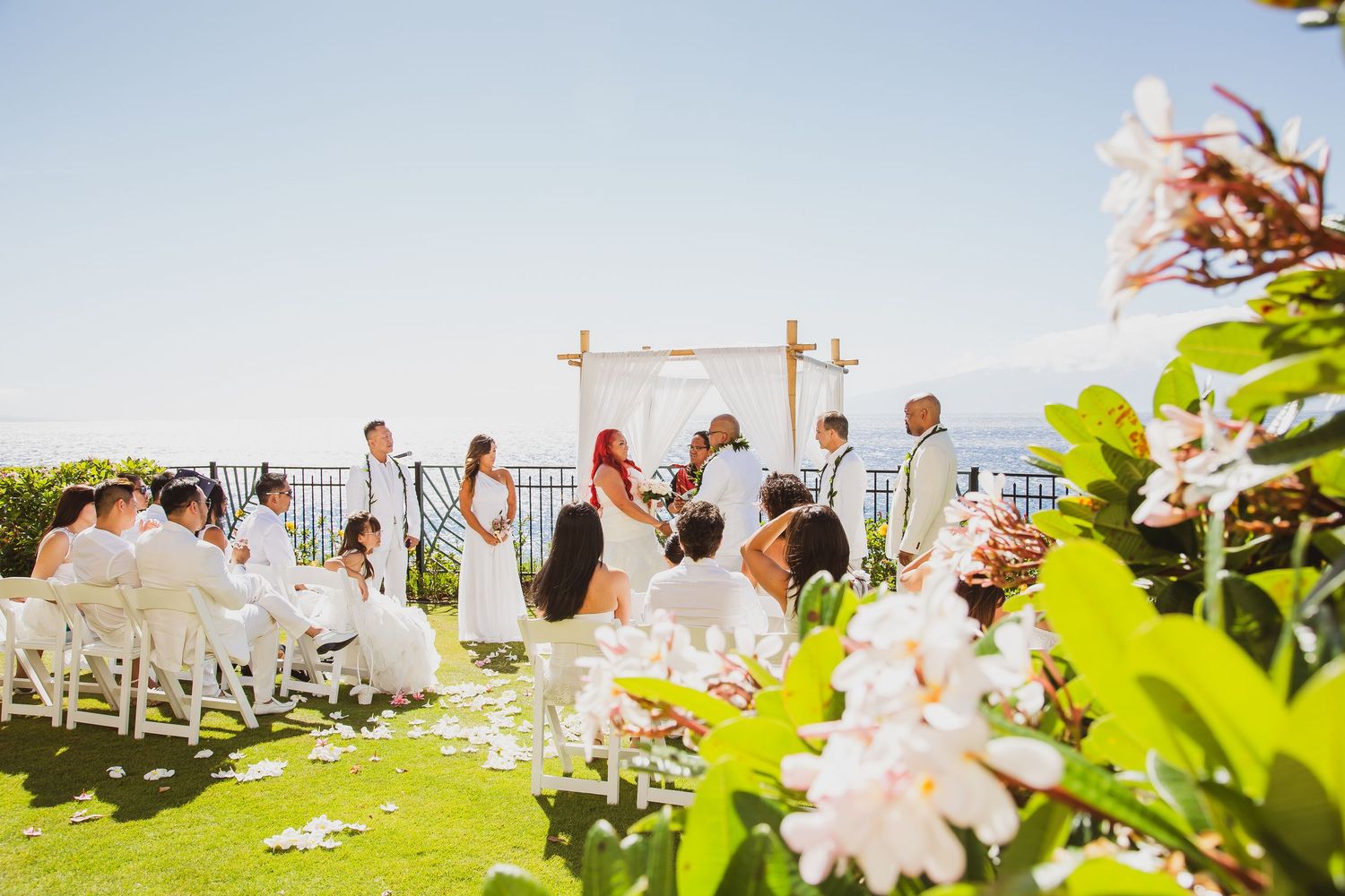 Outdoor wedding ceremony on a lush green lawn with ocean views, white chairs, floral arch, and tropical flowers in foreground.