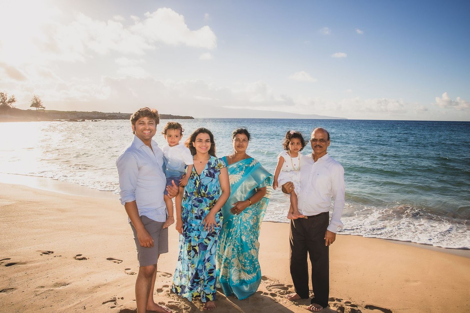 A group of people in casual and tropical attire pose together on a sunny beach with ocean waves in the background.