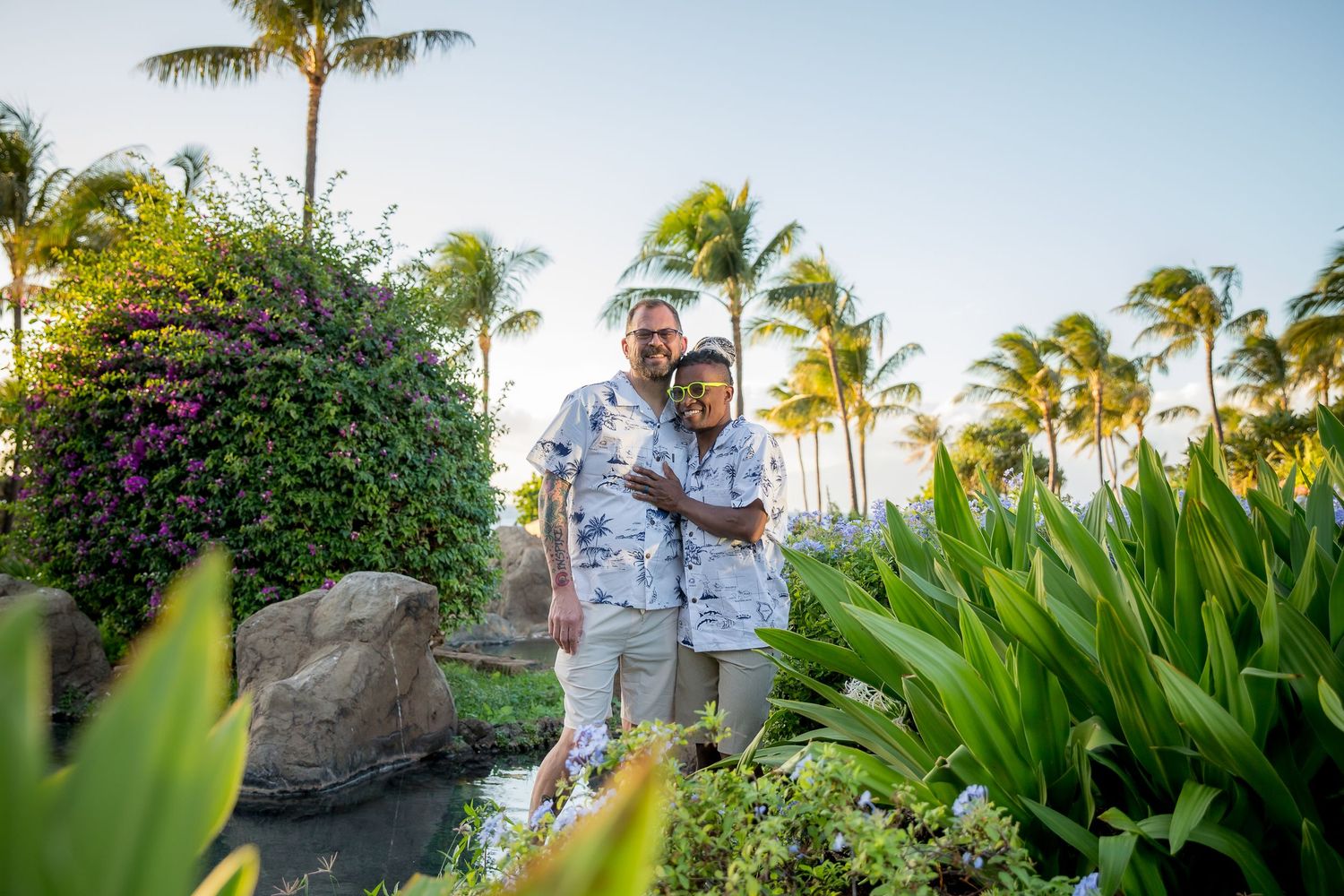 A couple stands in a tropical garden setting with palm trees and lush greenery at sunset in Hawaii.