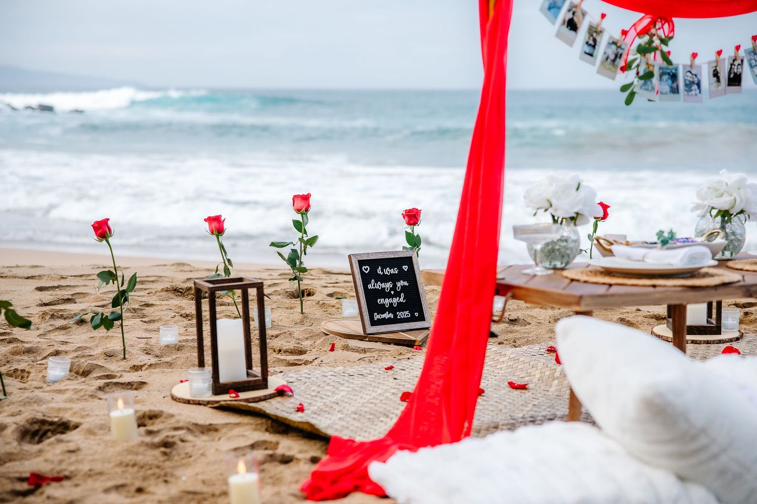 Red fabric decorations line a sandy beach with small signs and tropical flowers creating a romantic seaside setting.