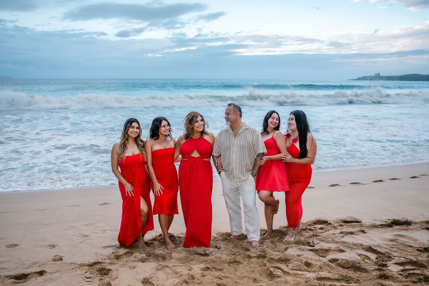Wedding party in flowing red dresses posing together on a sandy beach with ocean waves.