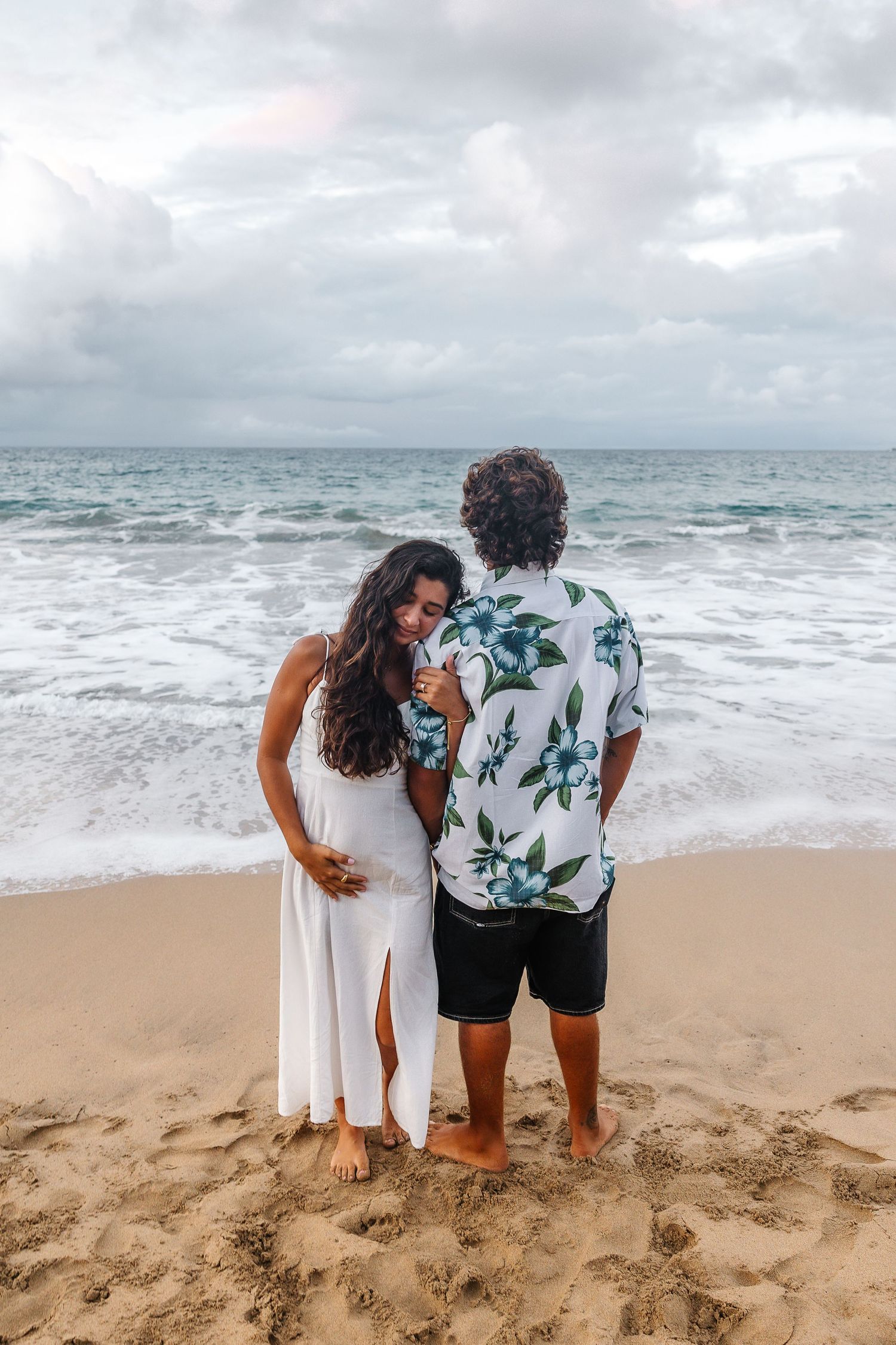 Couple stands together watching ocean waves on a cloudy beach day wearing coordinated outfits.