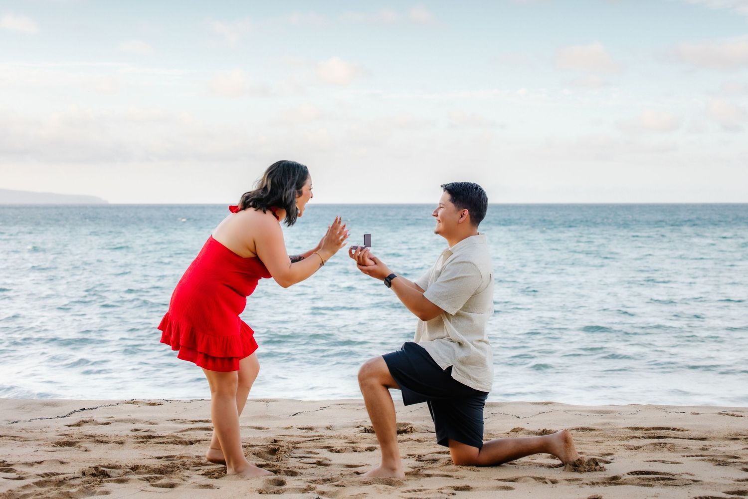 Man proposes to woman on sandy beach, she reacts with surprise as he kneels holding a ring box by the ocean.