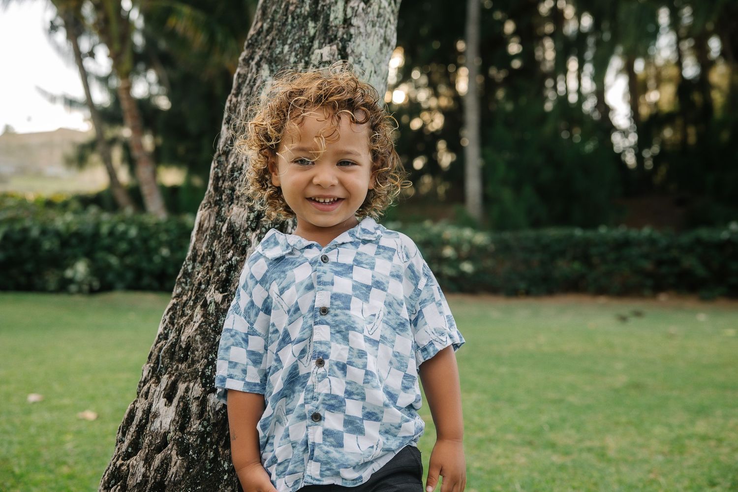 A child in a floral shirt plays outdoors on a sunny day in a grassy area near trees.