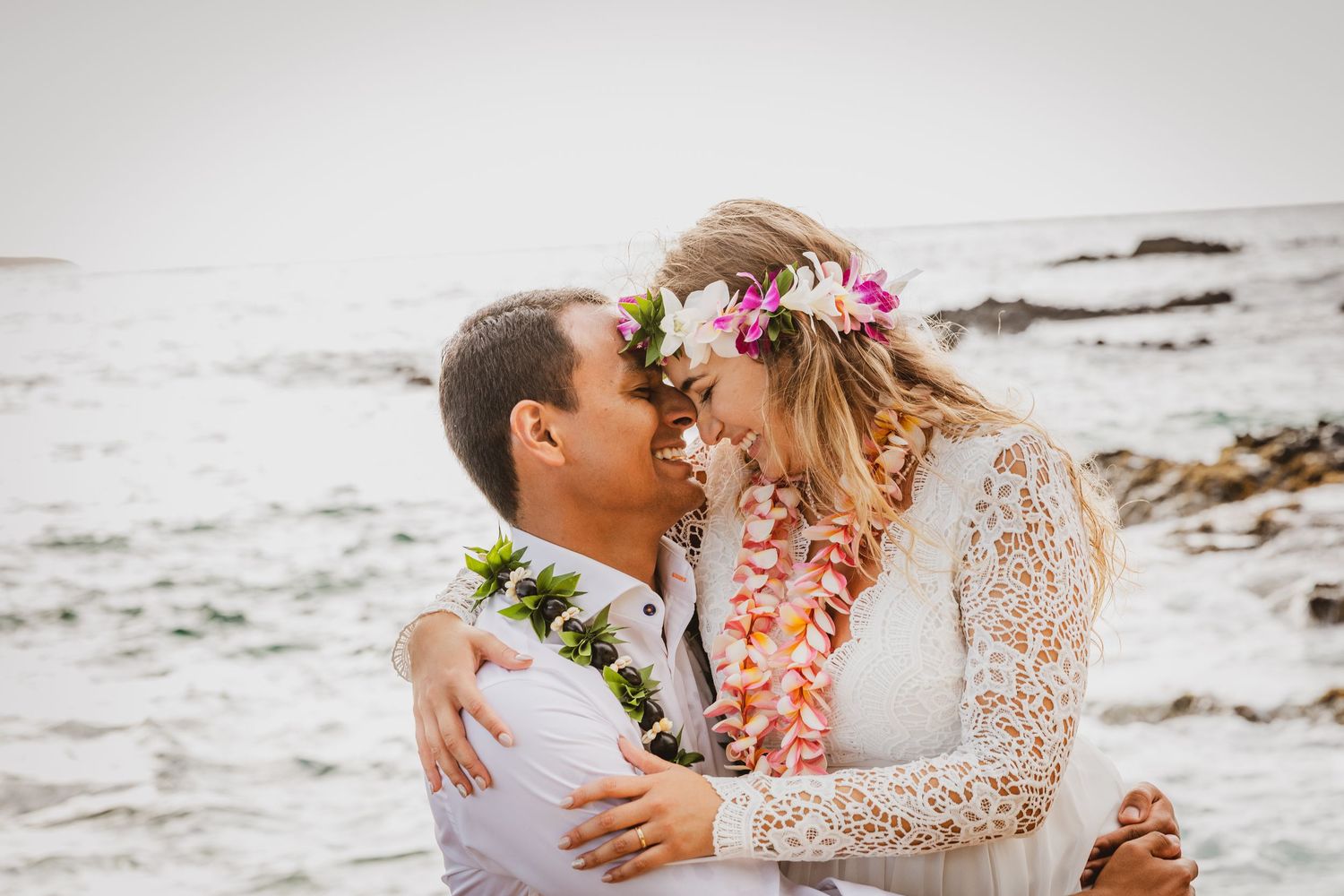 Couple sharing a romantic beach moment in Hawaiian leis and floral crown, with ocean waves in the background.