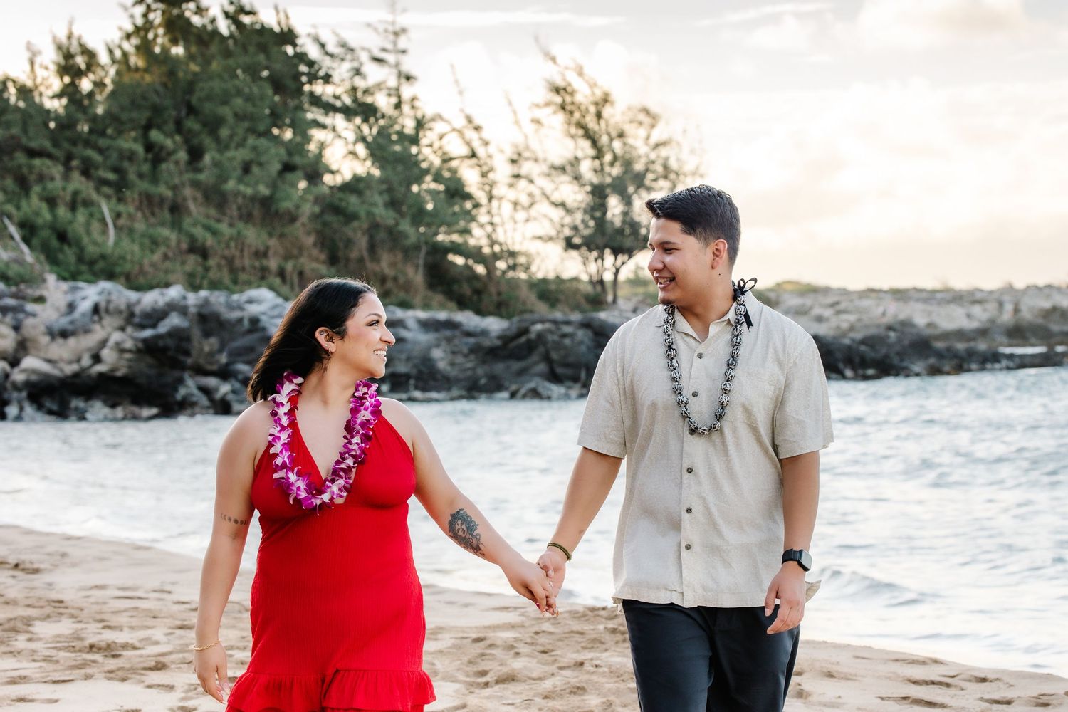 Couple holding hands and smiling on a Hawaiian beach at sunset, woman in red dress with lei, man in beige shirt.