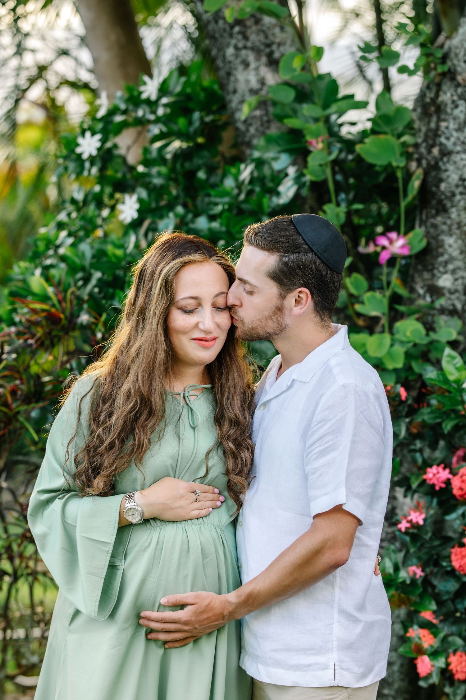 A man wearing a kippah kisses his pregnant wife's temple as they stand together in a lush tropical garden.